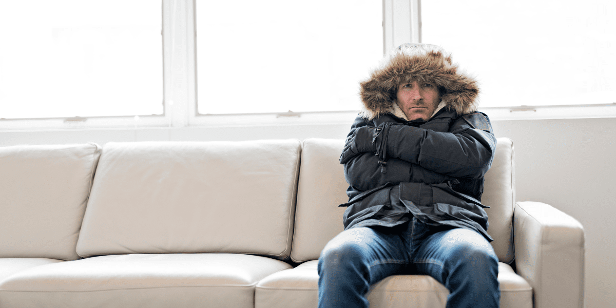 furnace blowing cold air in home, man wearing a winter coat sitting on couch